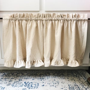 May include: A beige ruffled kitchen valance with a black rod hanging from white cabinets.