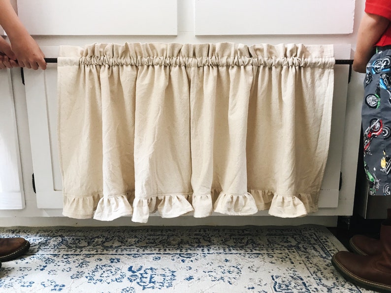 May include: A beige ruffled kitchen valance with a black rod hanging from white cabinets.