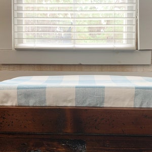 May include: A wooden bench with a blue and white checkered cushion. The bench is in front of a window with white blinds.