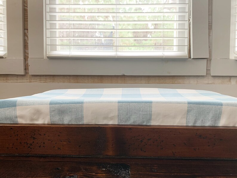 May include: A wooden bench with a blue and white checkered cushion. The bench is in front of a window with white blinds.