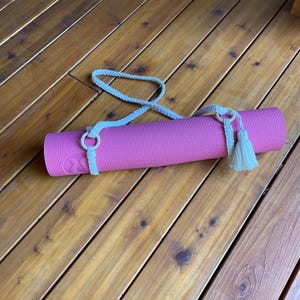 May include: A rolled-up pink yoga mat with a light blue strap and tassel, resting on a wooden deck. The strap is made of a woven material and is designed to carry the mat. The tassel is a light cream colour.
