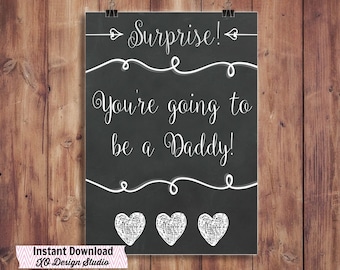 May include: A black chalkboard sign with white text that reads "Surprise! You're going to be a Daddy!" with three white hearts at the bottom. The sign is hanging on a wooden wall.