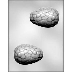May include: Two gray plastic molds for making chocolate eggs. Each mold is shaped like an egg with a textured surface that resembles a reptile's skin.