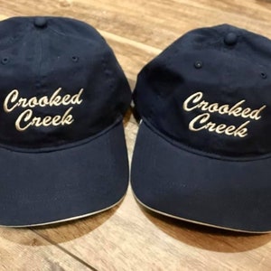 May include: Two navy blue baseball caps with a white brim. The caps have the text "Crooked Creek" embroidered in gold on the front.