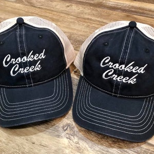 May include: Two navy blue and white mesh trucker hats with the text "Crooked Creek" embroidered in white on the front.