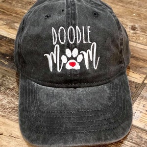 Op de afbeelding: Een zwarte, verweerde baseballpet met witte borduurwerk die "Doodle Mom" en een witte pootafdruk met een rood hartje zegt.