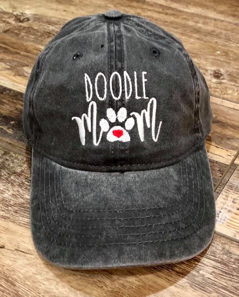Op de afbeelding: Een zwarte, verweerde baseballpet met witte borduurwerk die "Doodle Mom" en een witte pootafdruk met een rood hartje zegt.