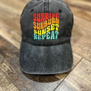 May include: Black baseball cap with a distressed look. The front of the cap features a colorful embroidered design that reads "Sunrise Sunburn Sunset Repeat".