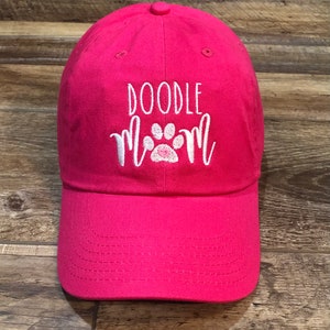 Op de afbeelding: Roze baseballpet met witte borduurwerk dat "Doodle Mom" zegt met een witte pootafdruk met een roze hart in het midden.