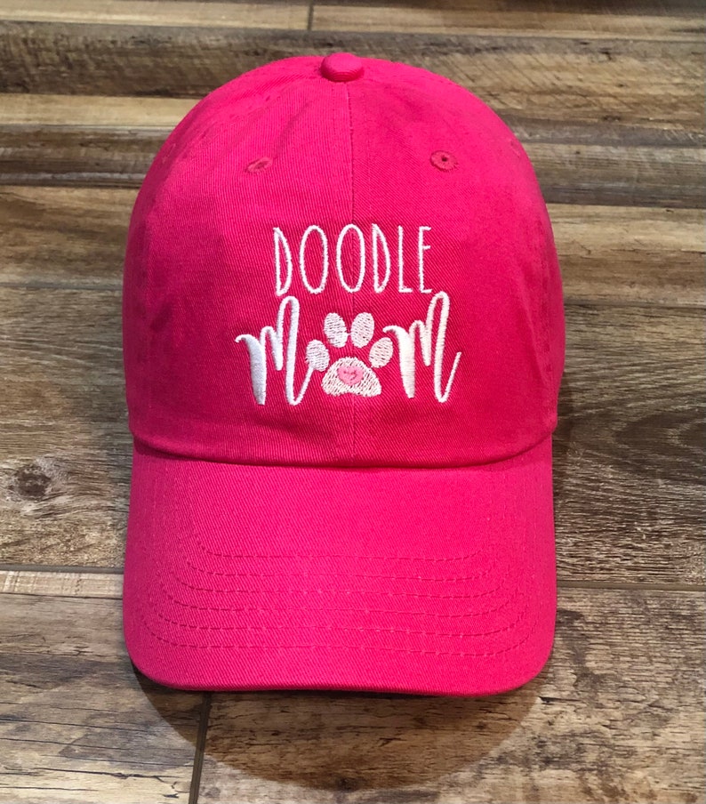 Op de afbeelding: Roze baseballpet met witte borduurwerk dat "Doodle Mom" zegt met een witte pootafdruk met een roze hart in het midden.