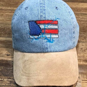 May include: Light blue denim baseball cap with a tan suede bill. The cap features an embroidered design of the state of Georgia in blue, set against a red and white striped background, with blue lightning bolt accents.