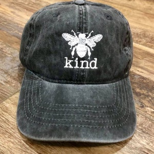 May include: A black distressed baseball cap with a white embroidered bee and the word "kind" underneath.