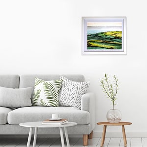 May include: A framed landscape photograph of rolling green hills with a body of water in the distance. The photograph is in a white frame and is hanging on a white wall.