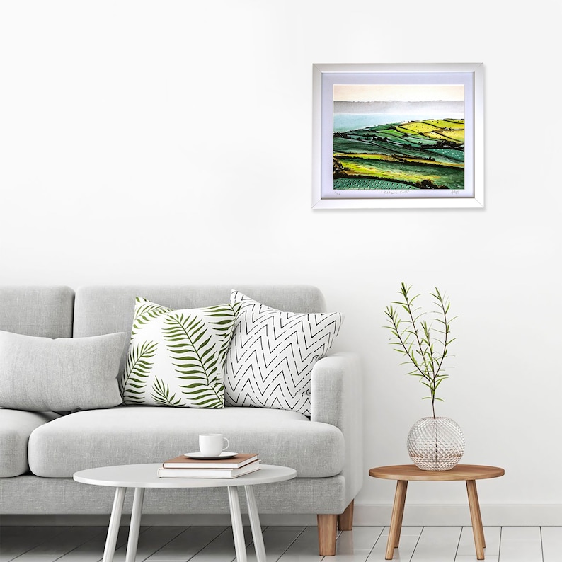 May include: A framed landscape photograph of rolling green hills with a body of water in the distance. The photograph is in a white frame and is hanging on a white wall.