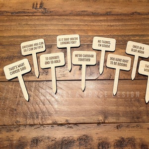 May include: Ten wooden plant markers with funny cheese puns. The markers are brown and have white text. The text on the markers includes: "Anything you can do I can do feta", "That's what cheese said", "To Havarti and to hold", "Is it brie you're looking for?", "We're cheddar together", "No thanks, I'm Gouda", "You have goat to be kidding", "Once in a blue moon", "Sweet dreams are made of cheese", and "Swiss you were here".