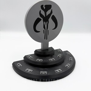May include: A gray and black Mandalorian symbol display. The circular emblem features a stylized skull design. The base is a tiered, black platform with small rectangular details. The text "JUST A TRACE" is visible.