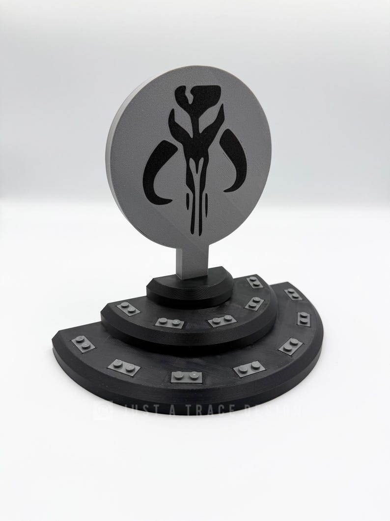 May include: A gray and black Mandalorian symbol display. The circular emblem features a stylized skull design. The base is a tiered, black platform with small rectangular details. The text "JUST A TRACE" is visible.