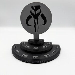May include: A black and gray 3D-printed Mandalorian symbol display. The design features a circular emblem with the Mandalorian skull symbol atop a tiered, semi-circular base. The base has small rectangular details. The text "JUST A TRACE DESIGN" is visible.
