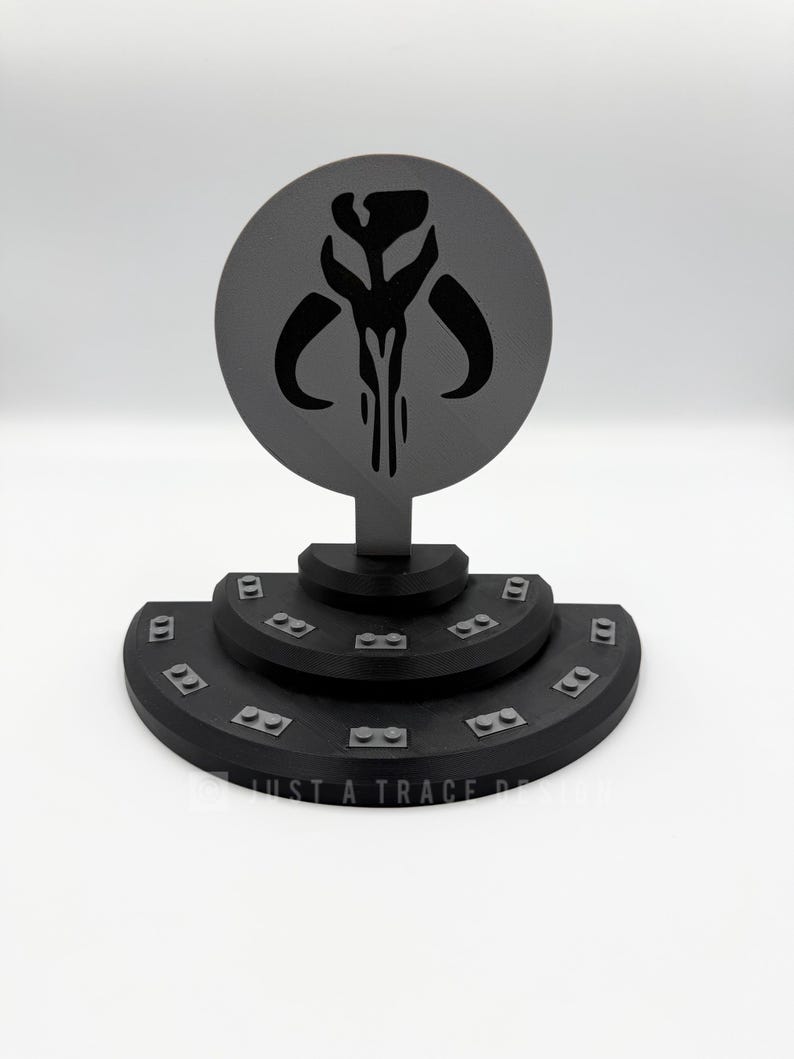 May include: A black and gray 3D-printed Mandalorian symbol display. The design features a circular emblem with the Mandalorian skull symbol atop a tiered, semi-circular base. The base has small rectangular details. The text "JUST A TRACE DESIGN" is visible.