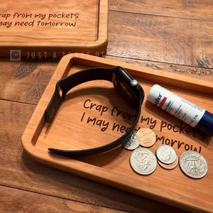 May include: Wooden tray with the engraved text "Crap from my pockets I may need tomorrow." The tray holds a black smartwatch, coins, and a white and blue glue stick. Another tray is visible in the background.