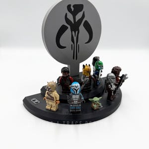 May include: A black display stand featuring several Star Wars Lego minifigures, including The Mandalorian and Grogu. A gray circular backdrop displays a Mandalorian symbol. The stand is on a white surface.