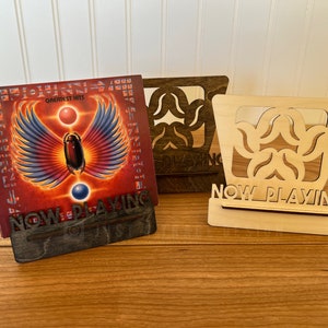 May include: Two wooden record stands with the words "Now Playing" on the front. The stand on the left is dark brown and the stand on the right is light brown. A vinyl record with a red, orange, and blue cover is displayed on the dark brown stand. The record cover features a winged creature and the words "Greatest Hits".