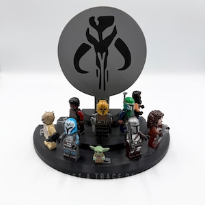 May include: A black display stand featuring several Star Wars Lego minifigures, including The Mandalorian and Grogu. A large, gray, circular backdrop displays a Mandalorian symbol. The base has the text "JUST A TRACE DESIGN".
