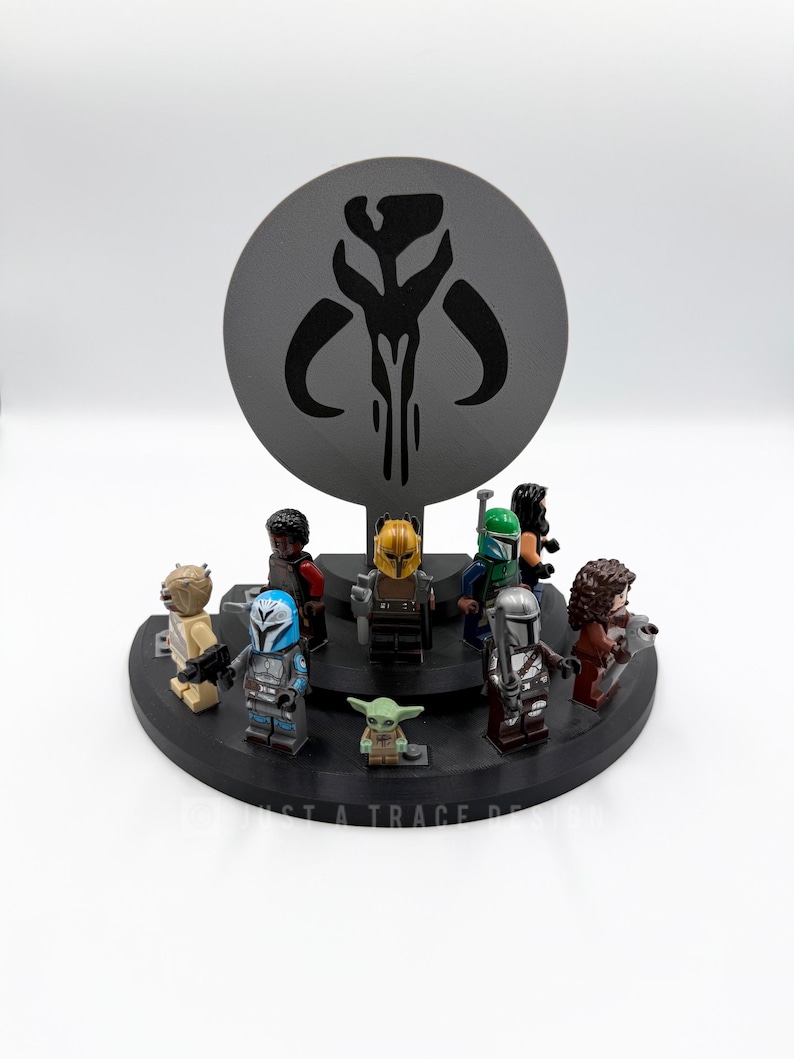 May include: A black display stand featuring several Star Wars Lego minifigures, including The Mandalorian and Grogu. A large, gray, circular backdrop displays a Mandalorian symbol. The base has the text "JUST A TRACE DESIGN".
