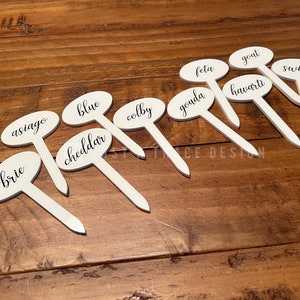 May include: White oval-shaped cheese markers with black lettering. The markers are arranged on a wooden surface. The cheese names are: brie, asiago, cheddar, blue, colby, gouda, feta, goat, havarti, and swiss.