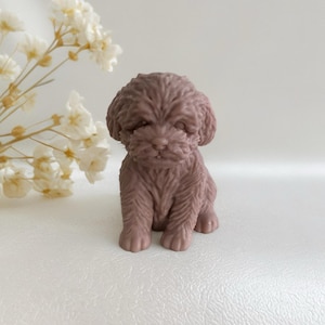 May include: A small, mauve-colored puppy figurine. The puppy is sitting upright with textured fur detail. The background is a soft white, with a sprig of white flowers in the upper left corner. The figurine is a decorative item.