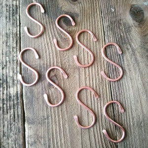 May include: Eight copper S-shaped hooks on a wooden surface. The hooks are all the same size and shape, and they are arranged in a random pattern.
