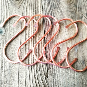 Peut inclure: Cinq crochets en forme de S en or rose, en m&eacute;tal. Les crochets sont dispos&eacute;s en rang&eacute;e sur une surface en bois.