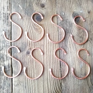 May include: Eight copper S-shaped hooks arranged in two rows of four on a wooden surface. The hooks are a warm, metallic copper colour.