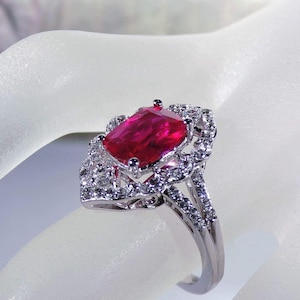 May include: A silver ring with a large, red, cushion-cut gemstone surrounded by smaller, clear gemstones.