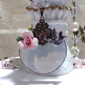 Peut inclure: Un collier en argent avec un pendentif rond en verre, une fleur rose et un motif d'oiseau. Le pendentif est d&eacute;cor&eacute; de strass roses. Le collier est sur un support blanc.