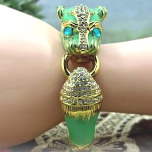 May include: A gold-toned bracelet featuring a green jaguar head with blue eyes and a green, jewel-encrusted pendant. The bracelet has a gold ring connecting the jaguar head and pendant. The bracelet is on a wrist.
