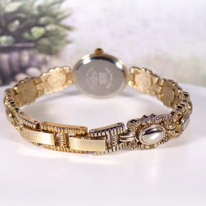 May include: Gold-toned wristwatch with an ornate, textured band. The watch face is round with a silver-colored back. The band features decorative oval and floral elements. The watch is displayed on a white surface.