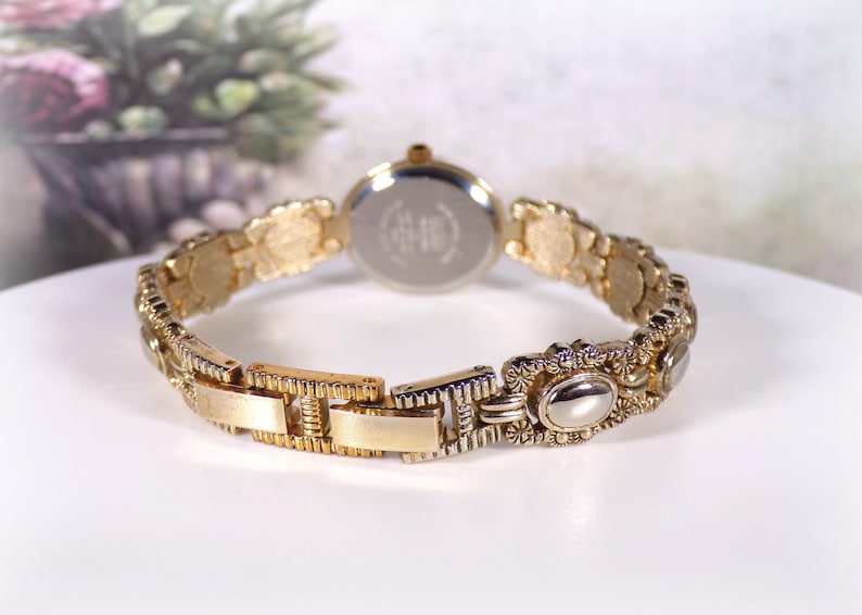 May include: Gold-toned wristwatch with an ornate, textured band. The watch face is round with a silver-colored back. The band features decorative oval and floral elements. The watch is displayed on a white surface.