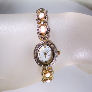 May include: A gold-toned wristwatch with a round face and a mother-of-pearl dial. The watch is adorned with clear crystals and pink oval-shaped stones. The band features decorative links with similar stones. The watch is a delicate piece of jewelry.