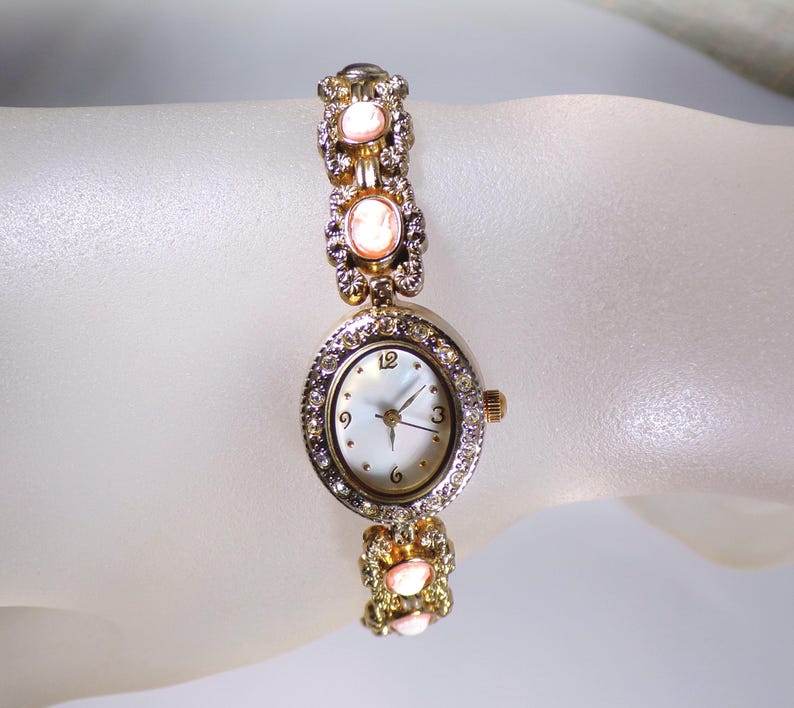 May include: A gold-toned wristwatch with a round face and a mother-of-pearl dial. The watch is adorned with clear crystals and pink oval-shaped stones. The band features decorative links with similar stones. The watch is a delicate piece of jewelry.