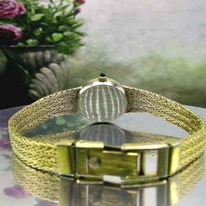 May include: A vintage gold-coloured wristwatch with a woven strap and a round face. The watch has a gold clasp and a small, dark-coloured stone on top. The watch is displayed on a reflective surface.
