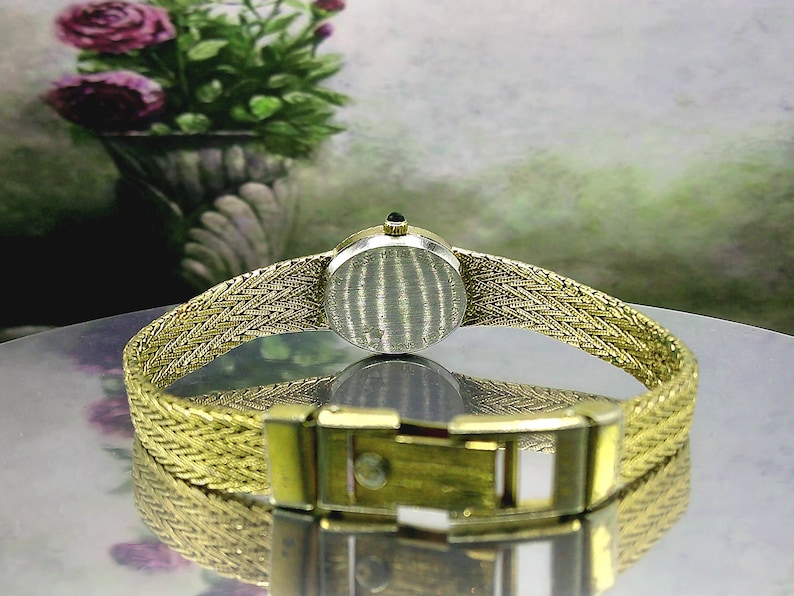 May include: A vintage gold-coloured wristwatch with a woven strap and a round face. The watch has a gold clasp and a small, dark-coloured stone on top. The watch is displayed on a reflective surface.
