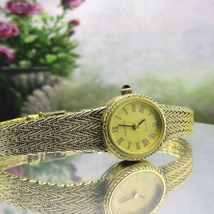 May include: A gold-coloured Movado quartz watch with a woven strap. The watch face displays Roman numerals and a black crown. The watch is reflected on a shiny surface. The background features a blurred floral arrangement.