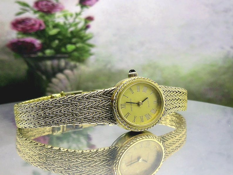 May include: A gold-coloured Movado quartz watch with a woven strap. The watch face displays Roman numerals and a black crown. The watch is reflected on a shiny surface. The background features a blurred floral arrangement.