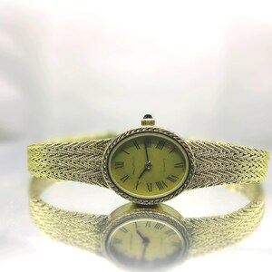 May include: A gold-coloured wristwatch with an oval face and a woven strap. The watch face features Roman numerals and a light green background. The strap and watch case are the same gold colour. The watch is reflected on a shiny surface.