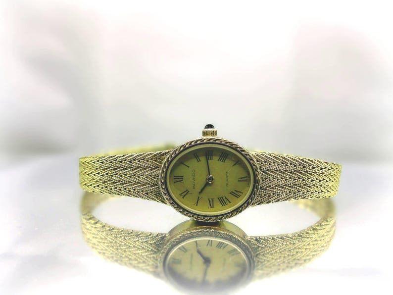 May include: A gold-coloured wristwatch with an oval face and a woven strap. The watch face features Roman numerals and a light green background. The strap and watch case are the same gold colour. The watch is reflected on a shiny surface.