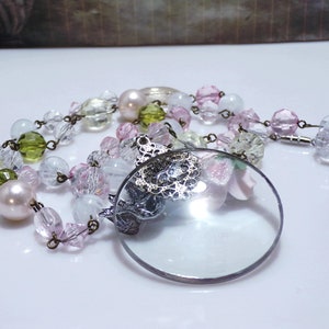 Peut inclure: Un collier vintage avec une loupe en verre, orn&eacute; d'un motif en filigrane argent&eacute; et d'un charme en forme de fleur rose. Le collier est compos&eacute; de perles de diff&eacute;rentes couleurs, notamment transparentes, roses, vertes et blanches.