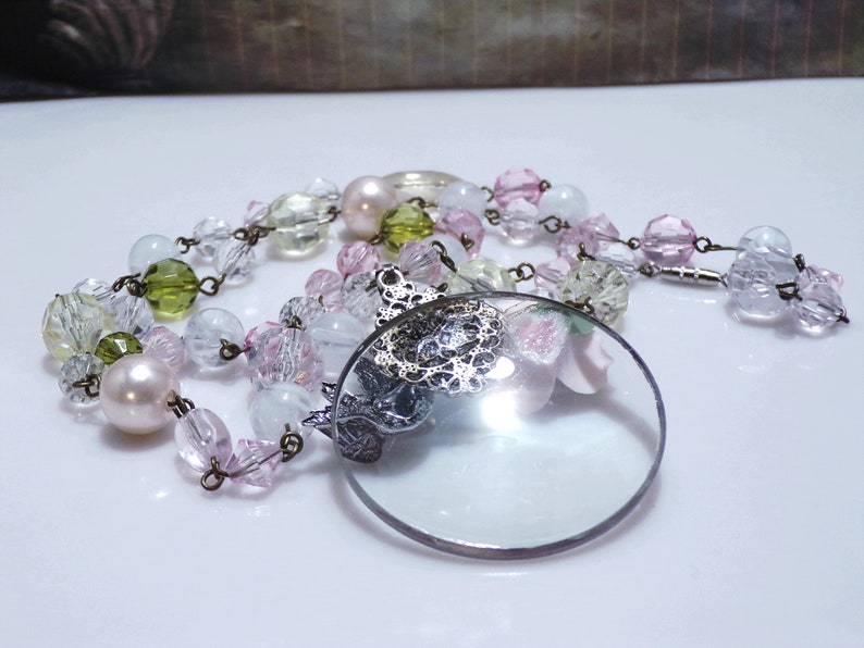 Peut inclure: Un collier vintage avec une loupe en verre, orn&eacute; d'un motif en filigrane argent&eacute; et d'un charme en forme de fleur rose. Le collier est compos&eacute; de perles de diff&eacute;rentes couleurs, notamment transparentes, roses, vertes et blanches.