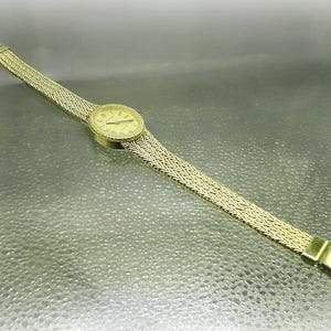 May include: Gold-coloured wristwatch with a woven strap. The watch face is round with Roman numerals. The strap is approximately 1.3 cm wide. The watch is set against a reflective surface.