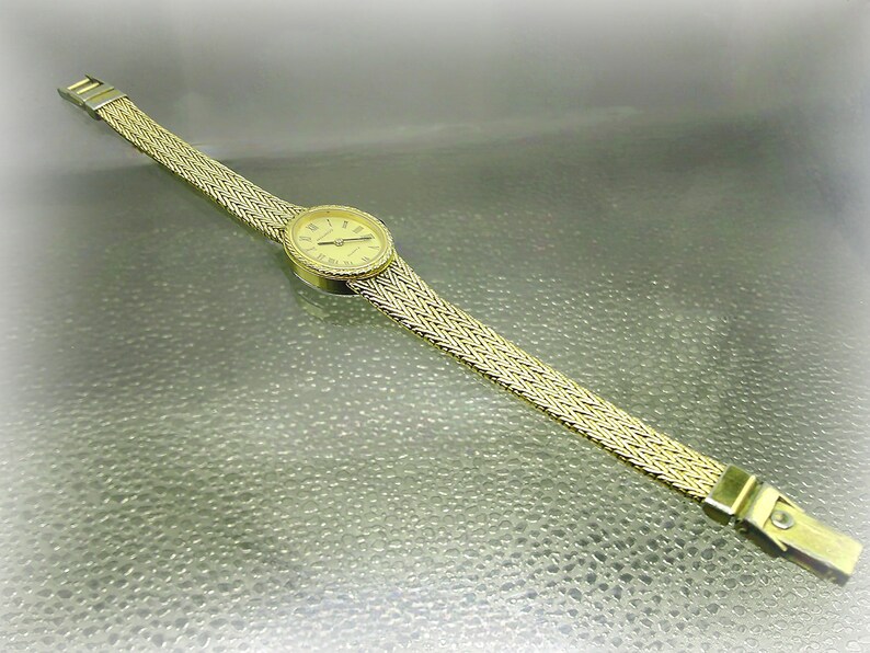 May include: Gold-coloured wristwatch with a woven strap. The watch face is round with Roman numerals. The strap is approximately 1.3 cm wide. The watch is set against a reflective surface.